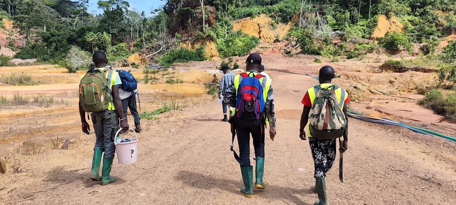 Trois personnes marchent vers un chantier d'exploitation, sacs au dos et bottes aux pieds.